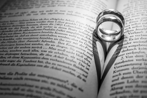 A pair of rings placed on an open book, casting a heart-shaped shadow on the pages. The text on the book is in black and white, with the focus centered on the rings and the shadow they create.
