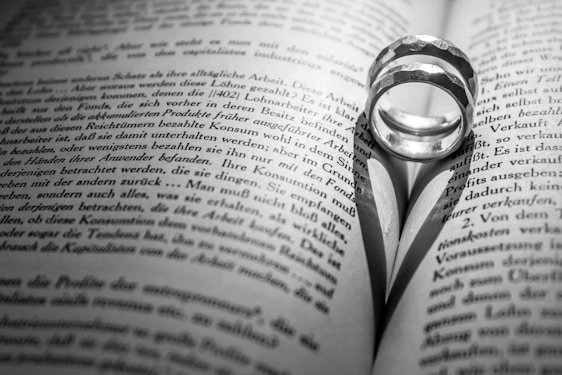 A pair of rings placed on an open book, casting a heart-shaped shadow on the pages. The text on the book is in black and white, with the focus centered on the rings and the shadow they create.