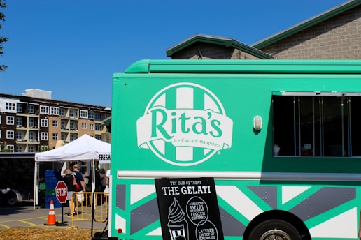 A green ice cream truck with a logo that reads 'Rita's Ice Custard Happiness' is parked in a market setting. The truck features promotional signs indicating their offerings, such as gelati and frozen custard. In the background, there is a white tent with people gathered around, possibly engaging in a market or outdoor event. Nearby buildings and a clear blue sky complete the scene.