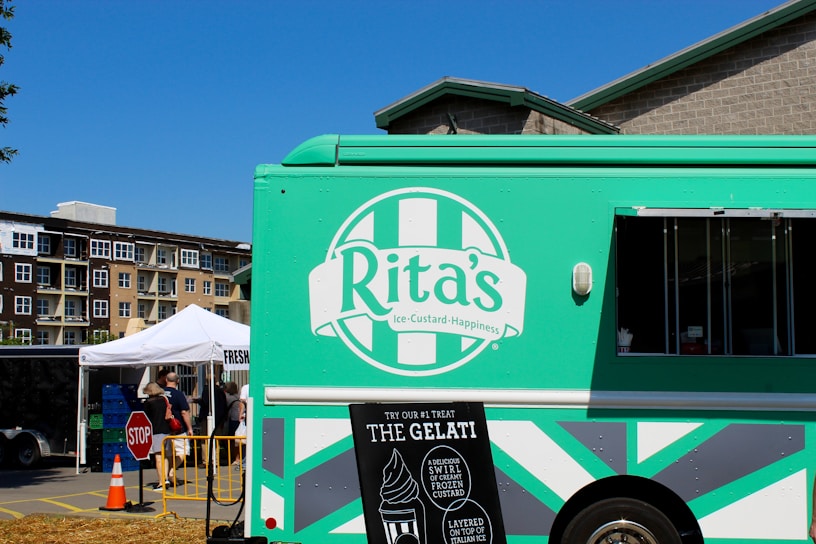A green ice cream truck with a logo that reads 'Rita's Ice Custard Happiness' is parked in a market setting. The truck features promotional signs indicating their offerings, such as gelati and frozen custard. In the background, there is a white tent with people gathered around, possibly engaging in a market or outdoor event. Nearby buildings and a clear blue sky complete the scene.