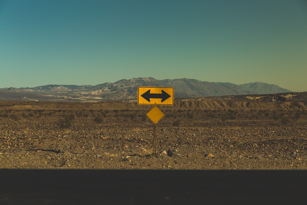 A yellow road sign with a black arrow pointing in both directions on the dry roadside near Death Valley