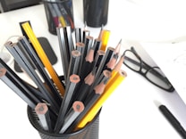 A collection of sharpened pencils, mostly black with some yellow ones, placed inside a mesh holder on a desk. Nearby are a pair of black glasses and a black pen. The background includes additional stationery items.