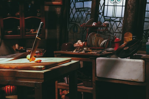 A rustic kitchen setting with dim lighting features a wooden table with a cutting board. A large knife is inserted into a sliced apple on the board. In the background, there are stained glass windows, shelves with various kitchen items, including dishes, utensils, and a dish rack containing plates.