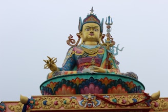 A large, colorful statue of a seated figure adorned with intricate ceremonial attire, including a headdress, necklaces, and a robe. The figure is holding objects like a trident and a ring. The base of the statue is decorated with ornate patterns and vibrant colors.