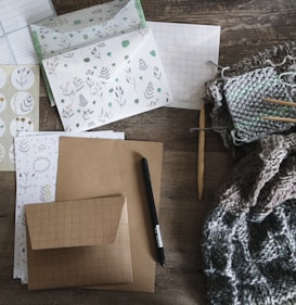 An assortment of stationery and knitting materials spread out on a textured wooden surface. Various items include patterned paper, envelopes with botanical designs, a black pen, and sheets with grid patterns. A knitting project is in progress, featuring yarn and knitting needles, which adds a cozy texture to the scene.