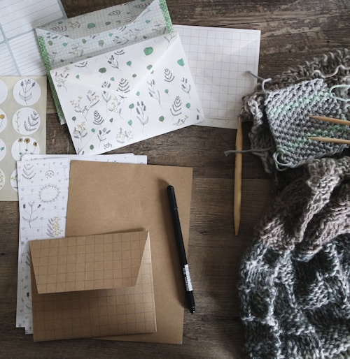 An assortment of stationery and knitting materials spread out on a textured wooden surface. Various items include patterned paper, envelopes with botanical designs, a black pen, and sheets with grid patterns. A knitting project is in progress, featuring yarn and knitting needles, which adds a cozy texture to the scene.