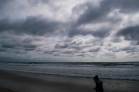Dark, overcast clouds hover above a vast ocean view, with waves gently reaching the shore. A person stands on the sand, possibly looking at a mobile device.