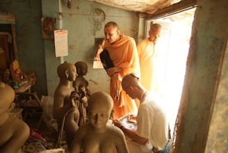 A group of people in a dimly lit room are surrounded by unfinished clay sculptures. Two individuals in traditional orange robes are observing the sculptures, while another person with a shaved head appears to be working on or studying one of the sculptures. The room has a rustic, authentic feel, with a worn-out calendar hanging on a blue wall and some religious artifacts visible in the background.