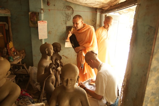 A group of people in a dimly lit room are surrounded by unfinished clay sculptures. Two individuals in traditional orange robes are observing the sculptures, while another person with a shaved head appears to be working on or studying one of the sculptures. The room has a rustic, authentic feel, with a worn-out calendar hanging on a blue wall and some religious artifacts visible in the background.