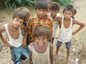 A group of children stand together outdoors, some with their arms around each other. They are on sandy ground with scattered plastic and foliage in the background. The children are dressed casually, some wearing vests and others in shorts and t-shirts.