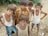 A group of children stand together outdoors, some with their arms around each other. They are on sandy ground with scattered plastic and foliage in the background. The children are dressed casually, some wearing vests and others in shorts and t-shirts.