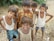 A group of children stand together outdoors, some with their arms around each other. They are on sandy ground with scattered plastic and foliage in the background. The children are dressed casually, some wearing vests and others in shorts and t-shirts.