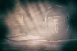An antique photograph of a man is enclosed in an oval frame, set against a hazy and obscured background. The dim lighting and soft focus give it a nostalgic and mysterious feel.