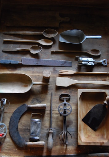 A collection of vintage kitchen utensils arranged on a wooden surface, including various spoons, scoops, and a grater. Among the items are an egg beater, a bread knife, a wooden fork, and a small tray. The tools exhibit signs of wear and rust, indicating their age and frequent use.