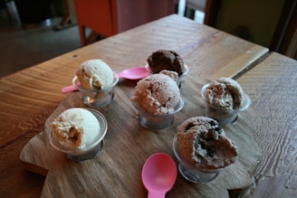 Six small cups of ice cream in various flavors are arranged on a wooden board. Two pink plastic spoons are placed among the cups. The ice cream flavors appear to range from vanilla to chocolate, with textures that look creamy and rich. The scene is set on a wooden table, giving a rustic feel to the composition.