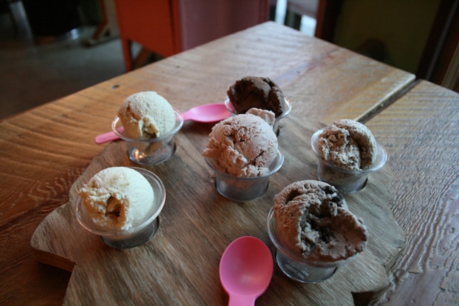 Six small cups of ice cream in various flavors are arranged on a wooden board. Two pink plastic spoons are placed among the cups. The ice cream flavors appear to range from vanilla to chocolate, with textures that look creamy and rich. The scene is set on a wooden table, giving a rustic feel to the composition.