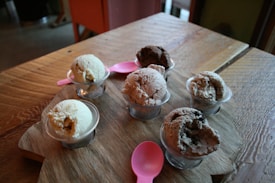 Six small cups of ice cream in various flavors are arranged on a wooden board. Two pink plastic spoons are placed among the cups. The ice cream flavors appear to range from vanilla to chocolate, with textures that look creamy and rich. The scene is set on a wooden table, giving a rustic feel to the composition.