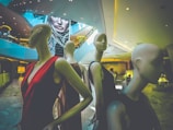 Mannequins dressed in elegant clothing are positioned within an upscale shopping mall or store interior. The space has modern architecture with sleek lines and a large digital display in the background featuring a woman's face. The ambiance is upscale and contemporary.