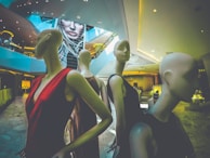 Mannequins dressed in elegant clothing are positioned within an upscale shopping mall or store interior. The space has modern architecture with sleek lines and a large digital display in the background featuring a woman's face. The ambiance is upscale and contemporary.