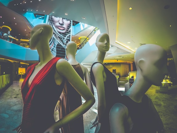 Mannequins dressed in elegant clothing are positioned within an upscale shopping mall or store interior. The space has modern architecture with sleek lines and a large digital display in the background featuring a woman's face. The ambiance is upscale and contemporary.