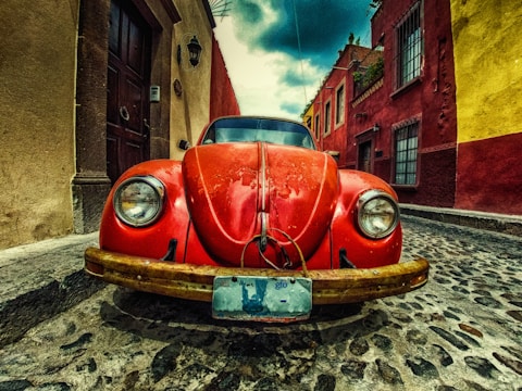 A vintage red Volkswagen Beetle is parked on a cobblestone street, surrounded by colorful colonial-style buildings in shades of yellow, red, and brown. The sky above is partly cloudy, adding a dramatic flair to the scene.