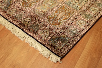 A richly detailed ornate Persian rug with intricate patterns in shades of orange, blue, and cream. The design includes floral motifs and geometric shapes, bordered with a fringe on one edge. Positioned on a wooden floor, the rug showcases traditional craftsmanship.