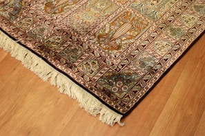 A richly detailed ornate Persian rug with intricate patterns in shades of orange, blue, and cream. The design includes floral motifs and geometric shapes, bordered with a fringe on one edge. Positioned on a wooden floor, the rug showcases traditional craftsmanship.