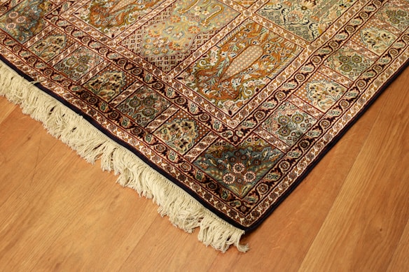 A richly detailed ornate Persian rug with intricate patterns in shades of orange, blue, and cream. The design includes floral motifs and geometric shapes, bordered with a fringe on one edge. Positioned on a wooden floor, the rug showcases traditional craftsmanship.