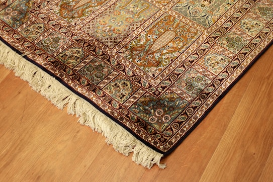 A richly detailed ornate Persian rug with intricate patterns in shades of orange, blue, and cream. The design includes floral motifs and geometric shapes, bordered with a fringe on one edge. Positioned on a wooden floor, the rug showcases traditional craftsmanship.