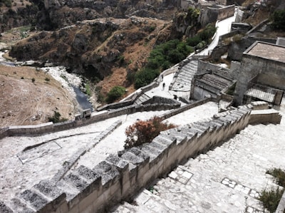 A series of stone steps and paths winding through a hilly landscape with rocky terrain and sparse vegetation. The scene features some ancient stone structures and retaining walls that line the pathways. A few people are visible walking along the paths, contributing to the sense of scale and human presence in the historical setting.