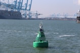 A large green buoy floating on the water with a solar panel attached to it. In the background, there are several large cranes and a massive container ship docked at a busy industrial port. The scene is set under a clear sky with sparse clouds.