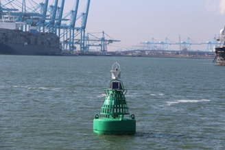 A large green buoy floating on the water with a solar panel attached to it. In the background, there are several large cranes and a massive container ship docked at a busy industrial port. The scene is set under a clear sky with sparse clouds.