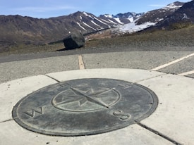A scenic view of a mountainous landscape with patches of snow on the peaks. In the foreground, there is a large, circular compass rose embedded in a concrete surface, marking the cardinal directions. A large rock is situated near the center, while the background displays a clear blue sky and rugged terrain.
