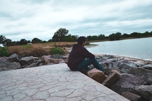 A solitary figure sitting by a calm lake, lost in thought under a vast sky.