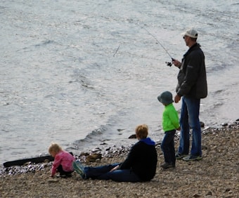 A man is fishing on a pebbly shore with a fishing rod, while two children and another person sit or stand nearby, engaging with the rocks and the water. The scene is peaceful, with calm water lapping at the shore.