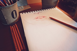 A sketchpad rests on a wooden table, featuring a partially completed drawing of lips in a pinkish hue. Multiple colored pencils lie beside the pad, with one placed directly on the pad, as if paused in the midst of drawing. A metal container wrapped in burlap holds more pencils, and the soft lighting casts warm shadows across the scene.