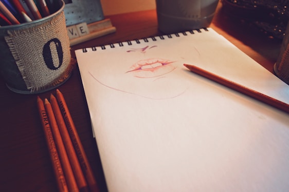 A sketchpad rests on a wooden table, featuring a partially completed drawing of lips in a pinkish hue. Multiple colored pencils lie beside the pad, with one placed directly on the pad, as if paused in the midst of drawing. A metal container wrapped in burlap holds more pencils, and the soft lighting casts warm shadows across the scene.