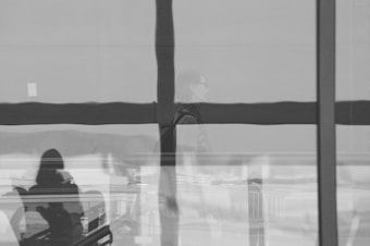 A black and white photograph captures reflections on a glass surface, showing silhouettes of people and elements of an interior space. The composition includes intersecting lines and reflections, creating an abstract and layered visual effect.