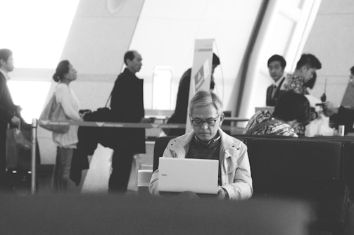 A black-and-white image depicting several people in an indoor setting, likely an airport or train station. In the foreground, a person is sitting on a bench using a laptop. In the background, a queue of people stands, some looking at the camera while others are engaged in conversation.