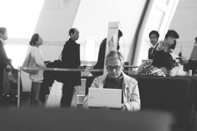 A black-and-white image depicting several people in an indoor setting, likely an airport or train station. In the foreground, a person is sitting on a bench using a laptop. In the background, a queue of people stands, some looking at the camera while others are engaged in conversation.