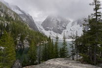 A serene lake is nestled among towering pine trees with a backdrop of snow-capped mountains. Misty clouds hover low, almost touching the mountain peaks, creating a tranquil and mysterious atmosphere.