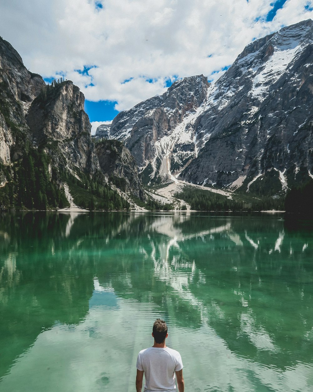 man standing in front of lake and cliff under white clouds and blue sky
