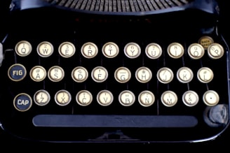 A close-up of an antique typewriter's keyboard featuring round, beige keys with black letters and numbers in a QWERTY layout. The surrounding frame is dark, with visible mechanical components near the top.