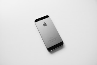 A black and silver smartphone, likely an older model iPhone, is placed on a plain white surface. The device is photographed from the back side, displaying the Apple logo prominently in the center.