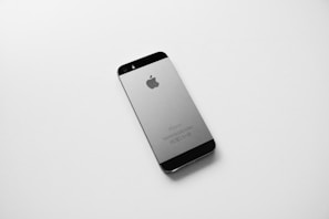 A black and silver smartphone, likely an older model iPhone, is placed on a plain white surface. The device is photographed from the back side, displaying the Apple logo prominently in the center.