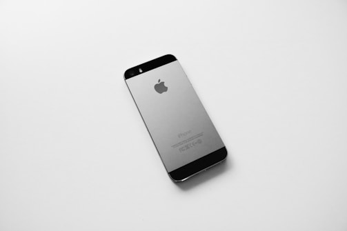 A black and silver smartphone, likely an older model iPhone, is placed on a plain white surface. The device is photographed from the back side, displaying the Apple logo prominently in the center.