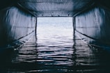 A tunnel-like structure with a corrugated metal roof and metallic walls frames a view of water with ripples, reflecting light from outside the opening.