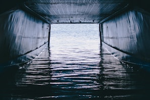A tunnel-like structure with a corrugated metal roof and metallic walls frames a view of water with ripples, reflecting light from outside the opening.