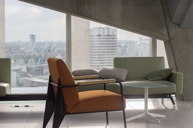 A modern interior scene with a focus on sleek furniture, including an orange armchair, a light green sofa, and a small round glass table. Large windows offer a view of a cityscape, allowing plenty of natural light to flood the space. The architecture features clean lines and concrete elements, contributing to a minimalist aesthetic.