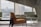 A modern interior scene with a focus on sleek furniture, including an orange armchair, a light green sofa, and a small round glass table. Large windows offer a view of a cityscape, allowing plenty of natural light to flood the space. The architecture features clean lines and concrete elements, contributing to a minimalist aesthetic.